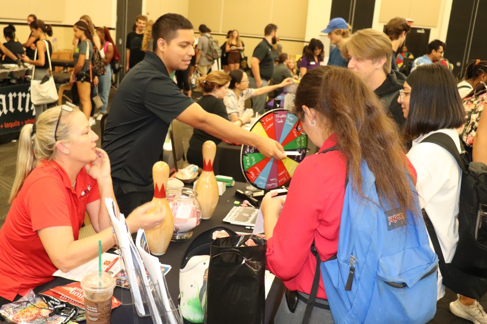 Students stand next to table with a prize wheel at the Welcome Expo while one person hands out a prize